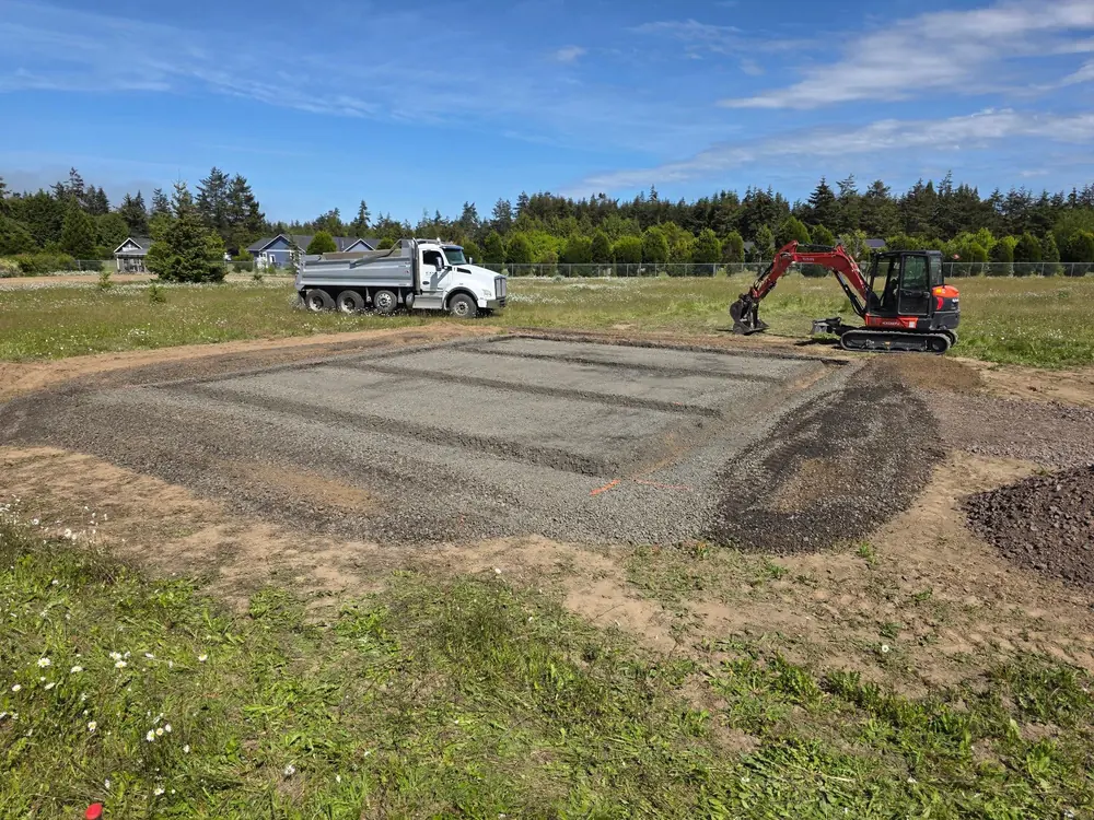 Excavator and dump truck shaping a compacted gravel building pad, by CKS Builders LLC in Port Angeles, WA