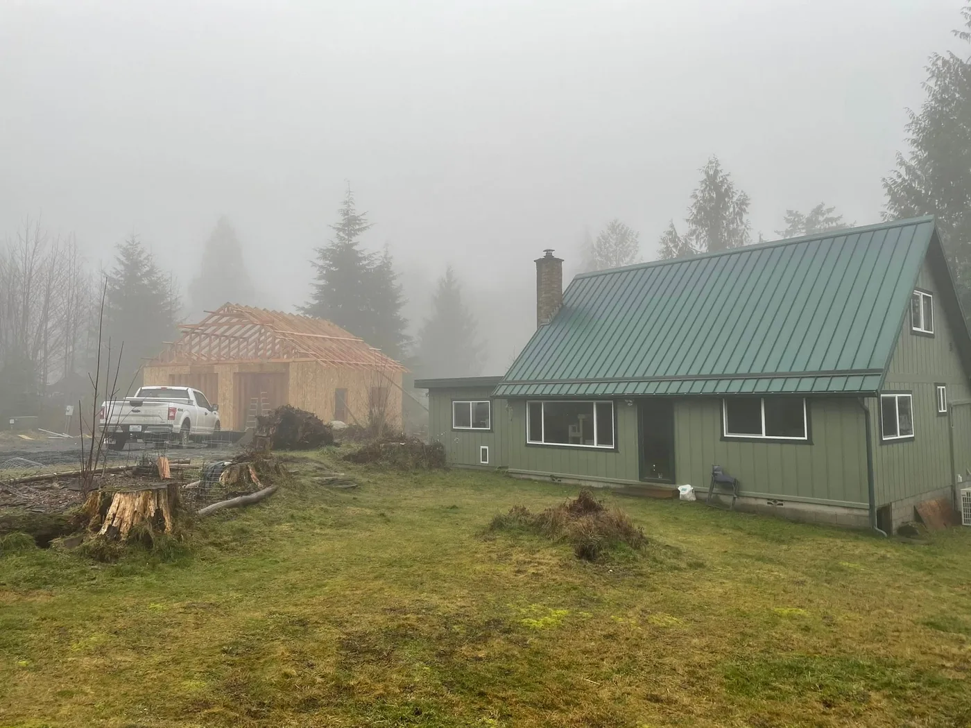 Small cottage with a green metal roof and a framed structure rising behind it on a foggy morning, by CKS Builders LLC in Port Angeles, WA